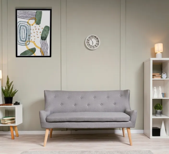 A gray tufted loveseat on tapered wooden legs, with framed abstract art and a wall clock above it and a white shelf to one side.
