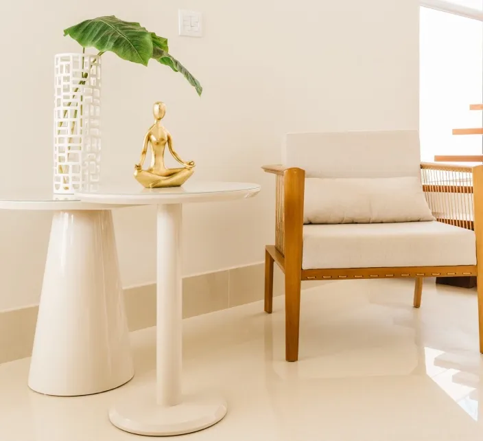 A cream pedestal side table holding a vase and a small gold figurine beside a wooden armchair with a white cushion.